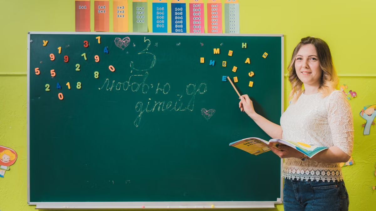 A female teacher pointing to a chalkboard with numbers in a colorful classroom.